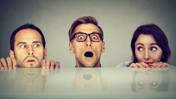 trzech ludzi wystawia głowę ponad stół, ten po środku robi przestraszoną minę